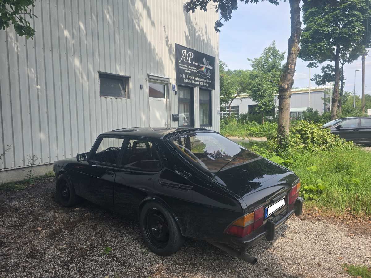 Ein schwarzer Saab 99 parkt vor einem Gebäude mit dem Schild „AP“. Sonniger Tag.
