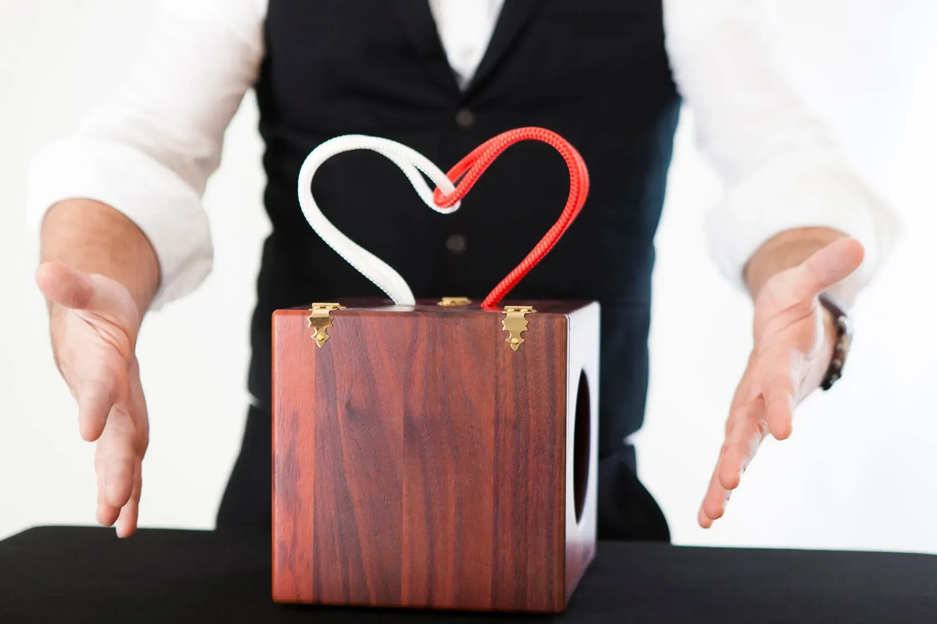 Hände umrahmen eine hölzerne Geschenkbox mit einem rot-weißen Herzband 