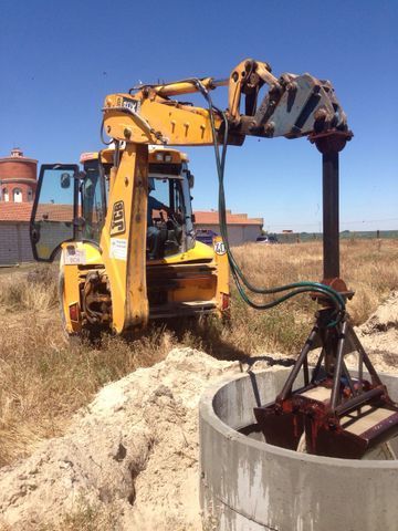 Excavadora amarilla levantando una plataforma de perforación junto a un pozo de hormigón en un campo de hierba bajo el cielo azul.
