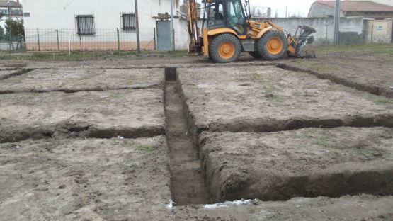 Retroexcavadora en una obra de construcción fangosa con zanjas profundas y un edificio al fondo.