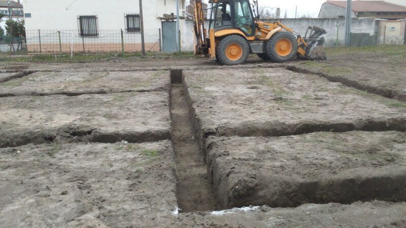 Obra en construcción con profundas zanjas en terreno fangoso y una retroexcavadora al fondo.