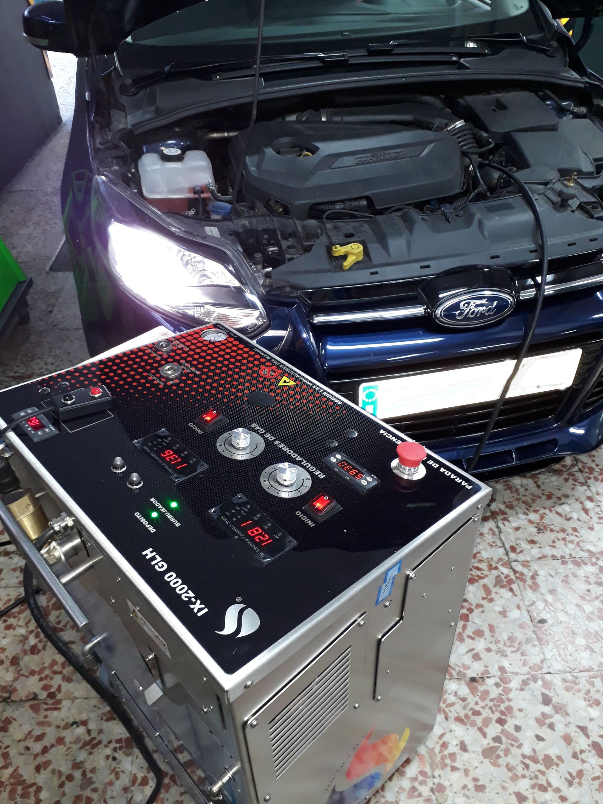 Una máquina especializada para la limpieza de carbono conectada al motor de un Ford azul oscuro estacionado en un taller mecánico.