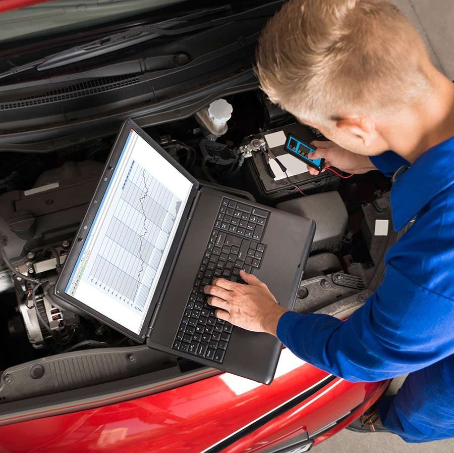 Un mecánico con uniforme azul utiliza un ordenador portátil y un dispositivo de mano para diagnosticar el motor de un coche bajo el capó abierto.