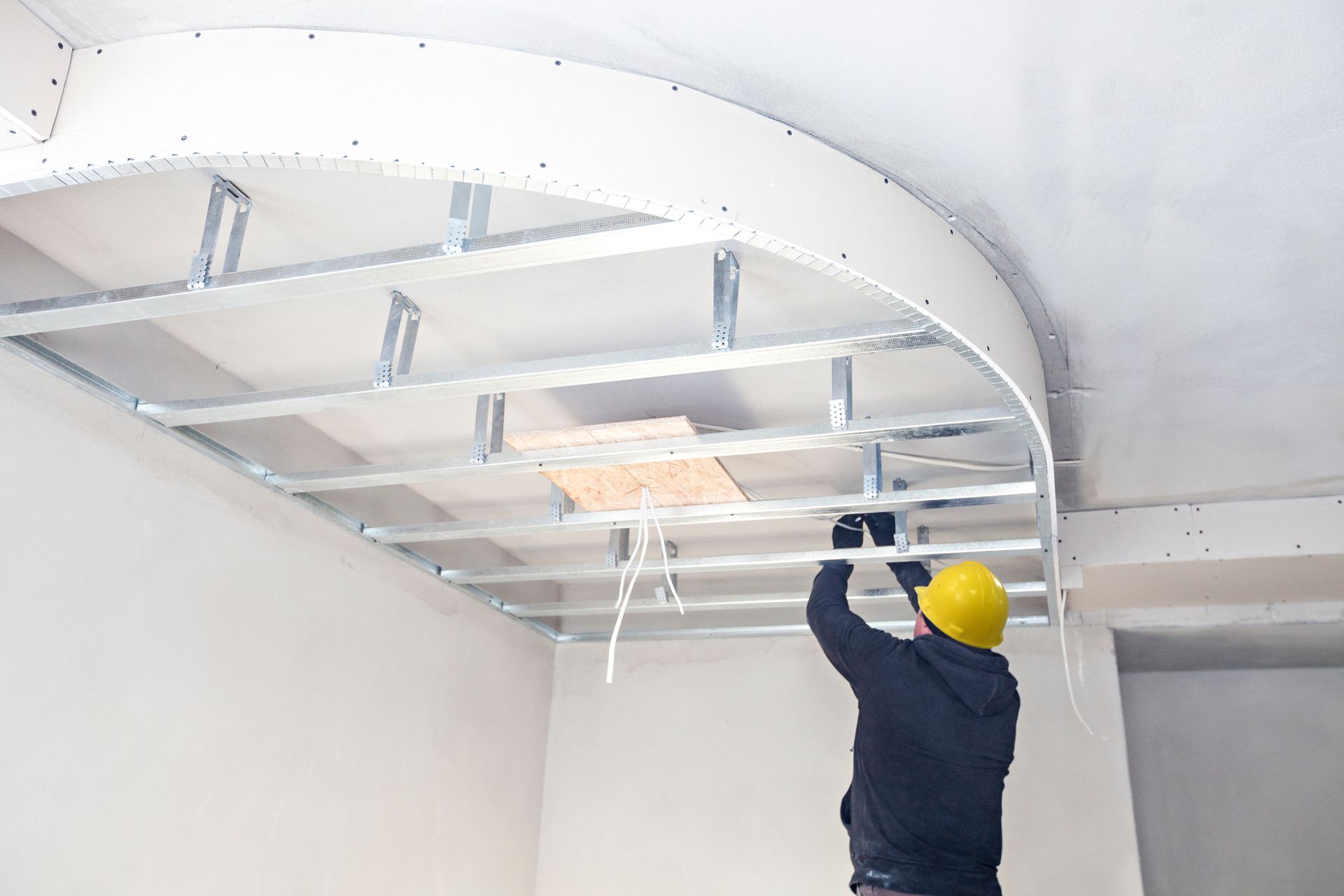 Ouvrier installant une ossature de plafond incurvée, portant un casque de chantier jaune, dans un intérieur blanc