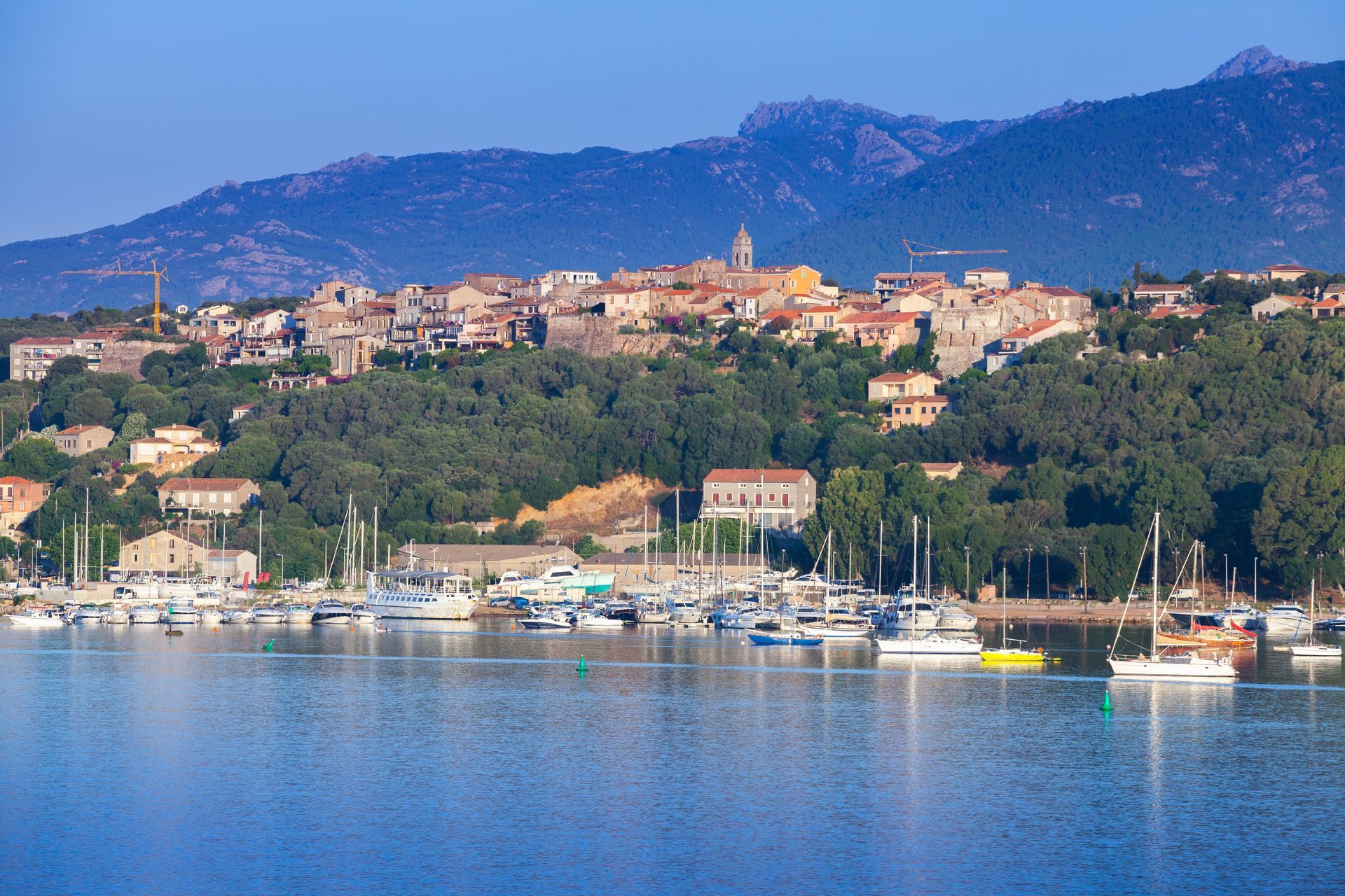 Ville côtière avec marina et voiliers, adossée à des collines verdoyantes et des montagnes sous un ciel d'un bleu limpide.
