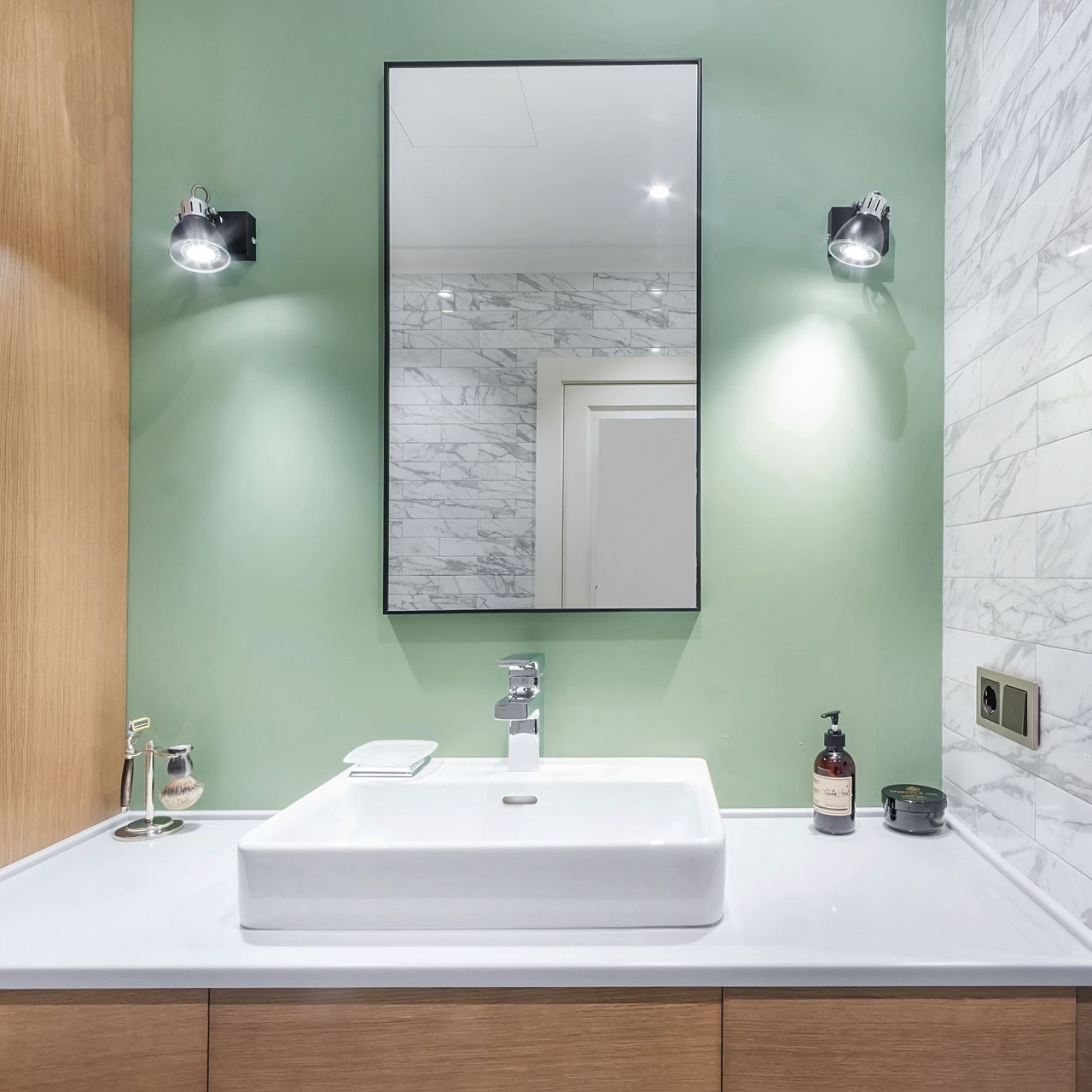 Meuble-lavabo moderne avec lavabo blanc, miroir à cadre noir et murs verts flanqués d'appliques murales.