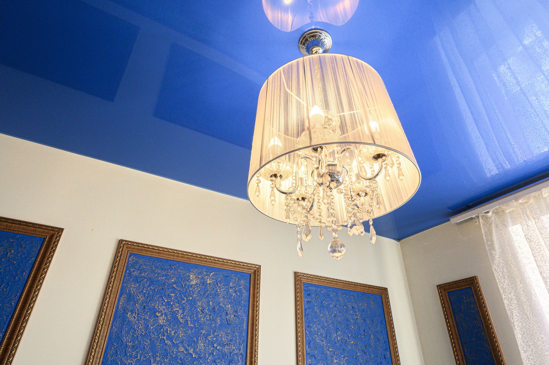 Un lustre en cristal est suspendu à un plafond bleu, au-dessus d'œuvres d'art bleues encadrées et de rideaux blancs.