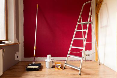 Pièce avec un mur rouge, une échelle, un rouleau à peinture et du matériel de peinture sur un sol en bois