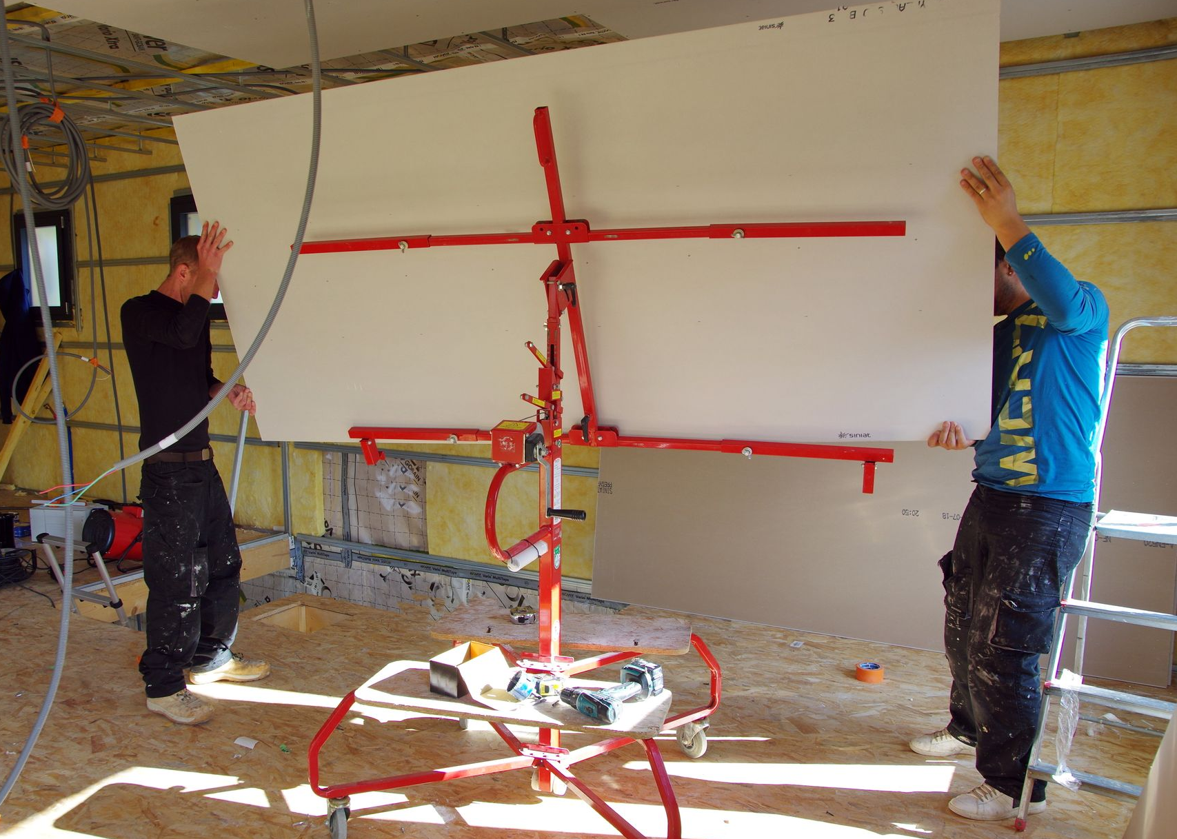 Deux ouvriers utilisent des ventouses rouges pour positionner un grand panneau mural blanc dans une pièce en cours de finition.