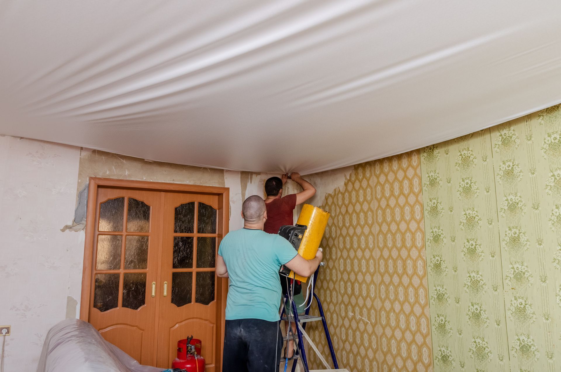 Deux personnes installent de l'isolant au plafond d'une petite pièce équipée d'une échelle et de portes en bois.