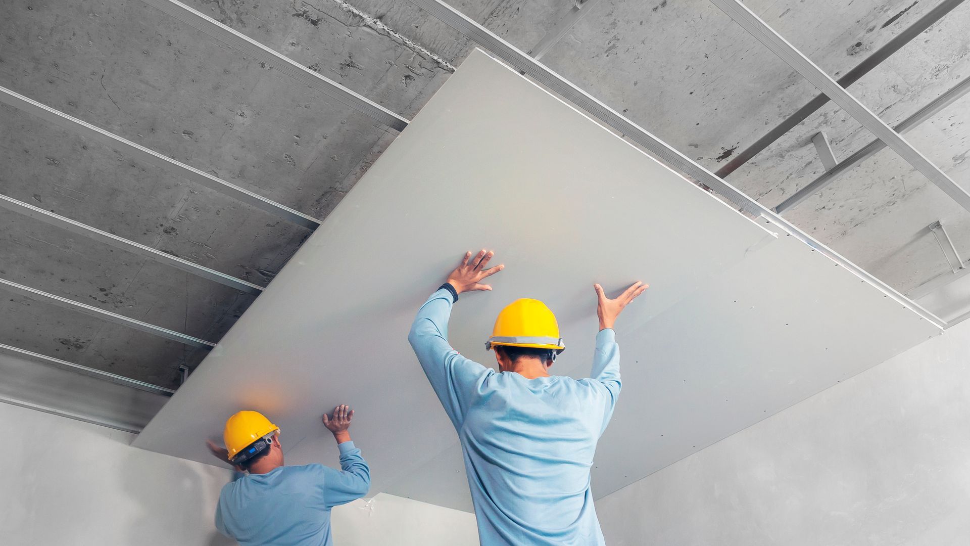 Deux ouvriers du bâtiment portant des casques jaunes installent un grand panneau de plafond blanc à l'intérieur.