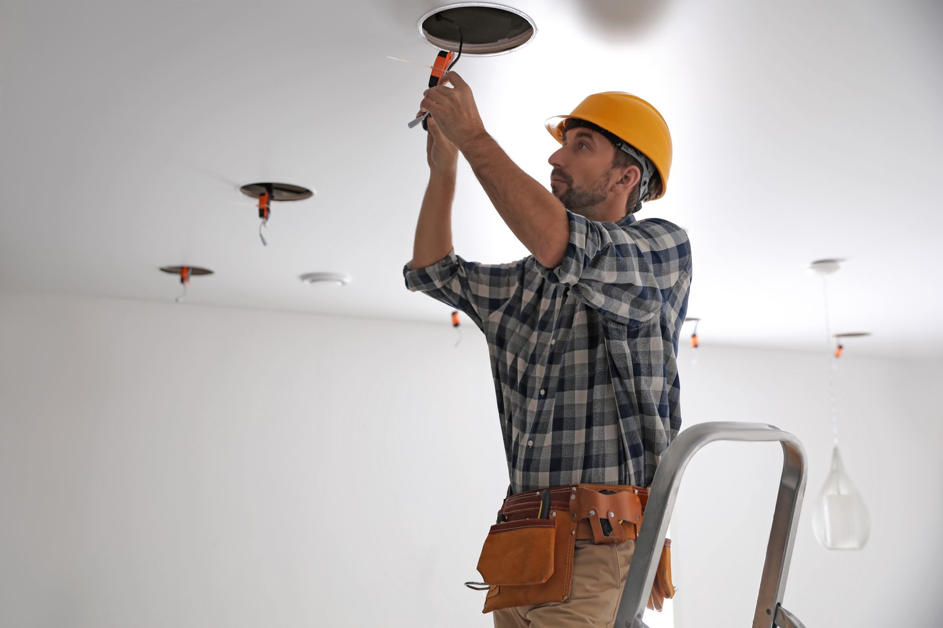 Un électricien portant un casque de chantier jaune installe un plafonnier dans une pièce blanche.