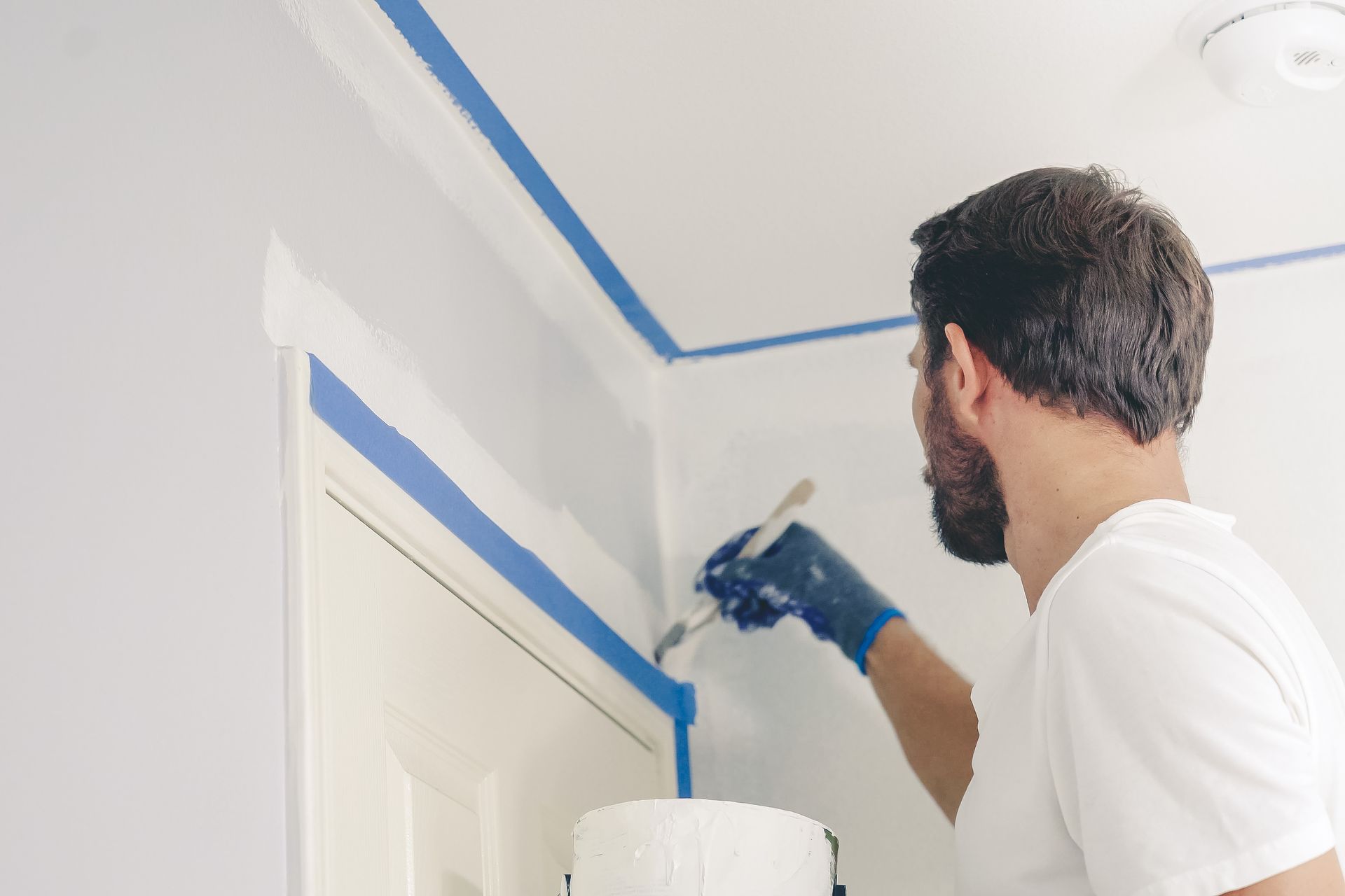 Un peintre applique du ruban adhésif bleu le long d'un angle de mur et de plafond blanc à l'intérieur.