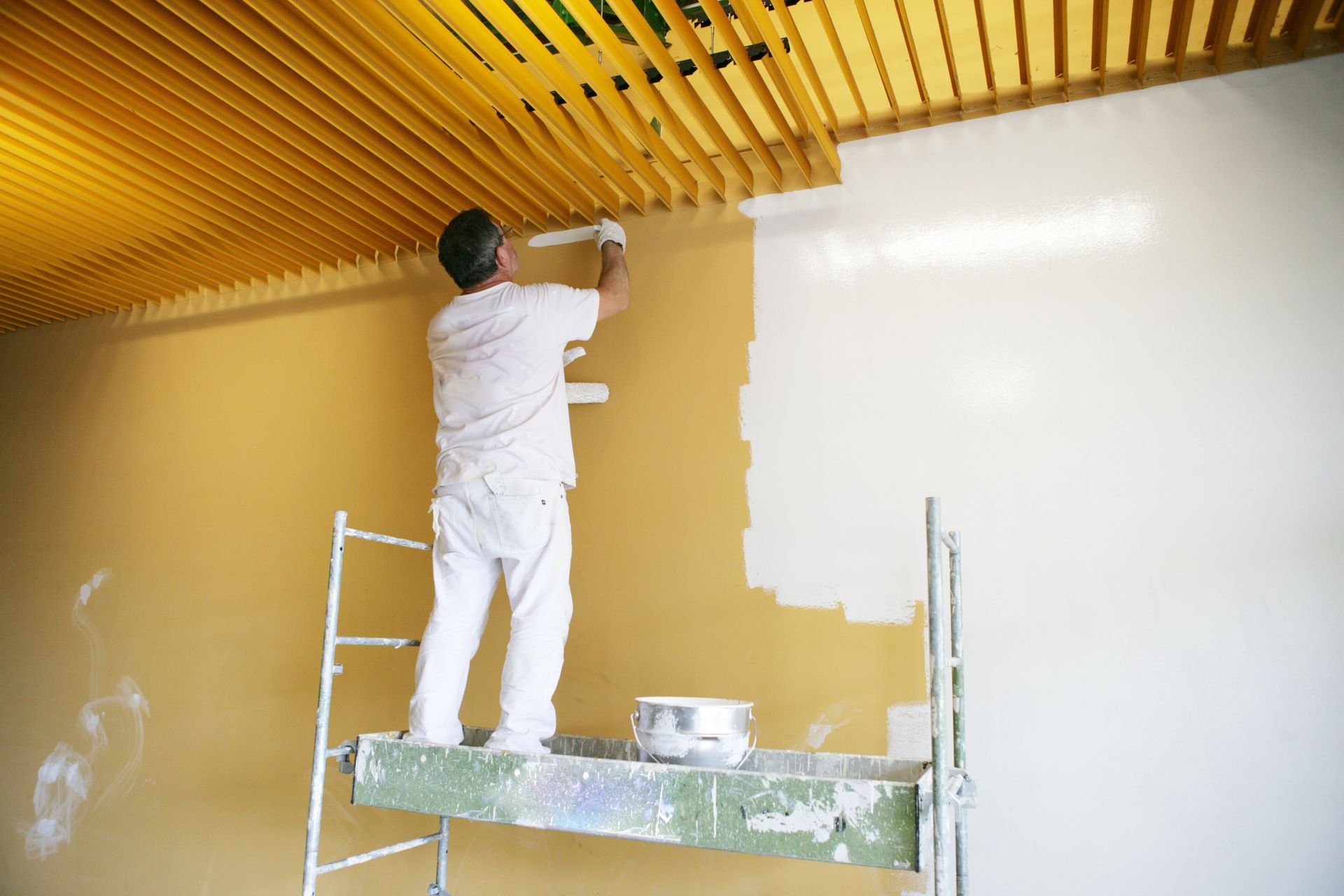 Une personne peint un mur jaune depuis un échafaudage, avec une partie peinte en blanc sur la droite.