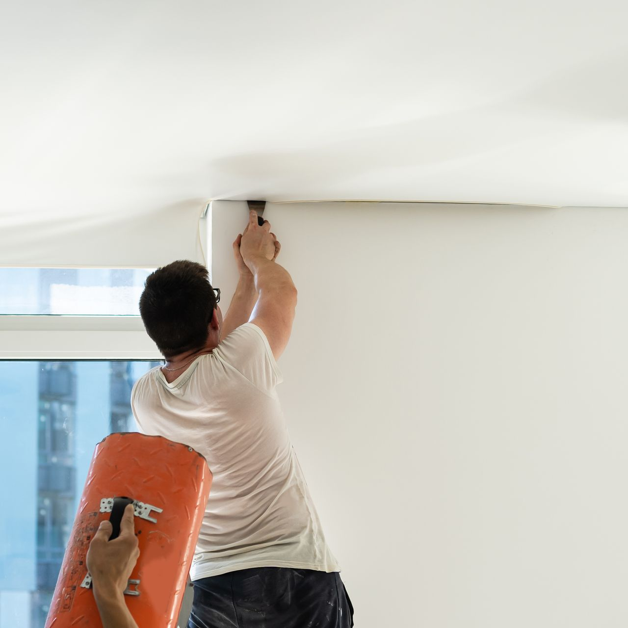 Une personne utilise une perceuse pour installer un luminaire sur un plafond blanc près d'une fenêtre.