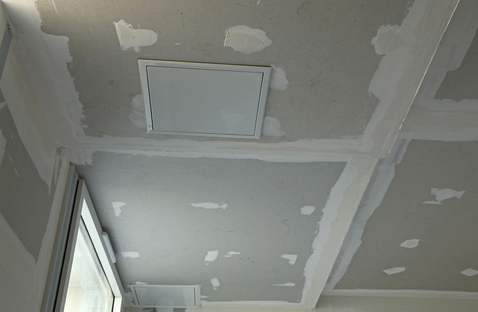 Plafond inachevé avec des réparations de plaques de plâtre, des joints et une trappe d'accès encastrée près d'une fenêtre.
