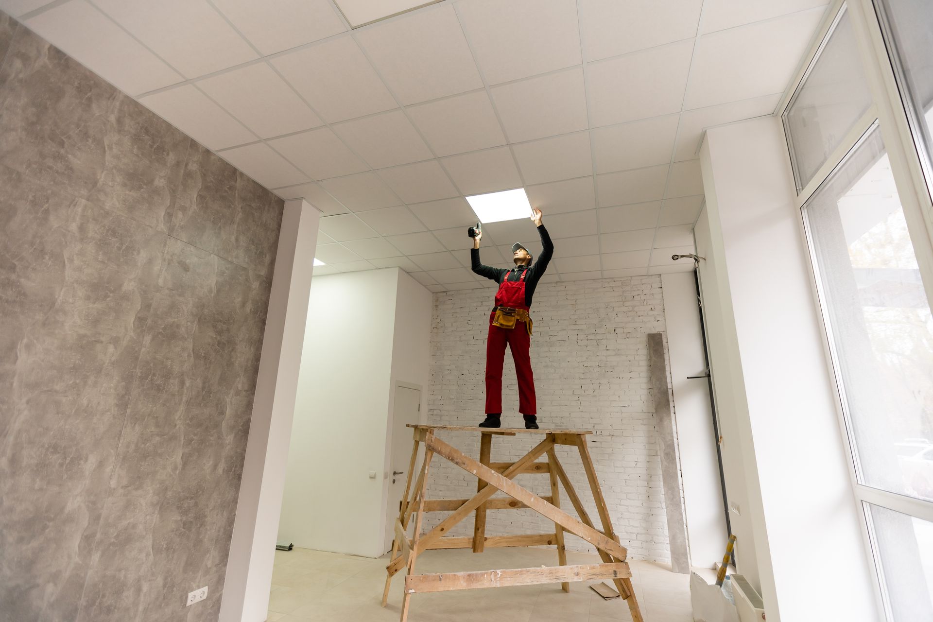 Un ouvrier, debout sur un échafaudage en bois, tient un panneau lumineux de plafond dans une pièce en travaux.