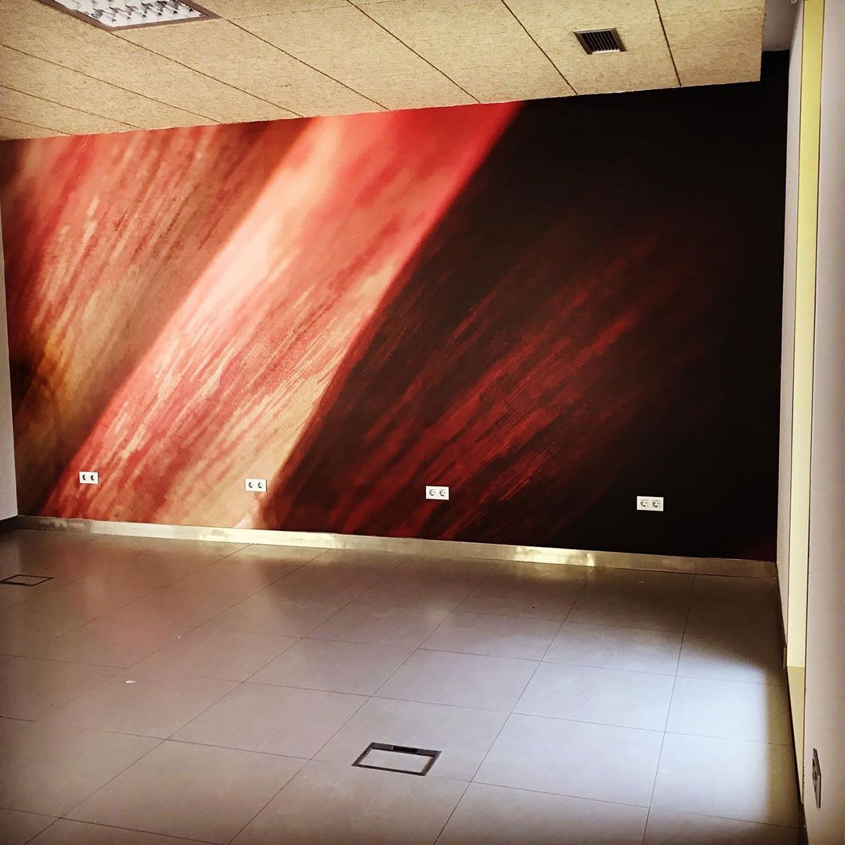 Habitación vacía con mural de color marrón rojizo en la pared, luz brillante y sombras, suelo de baldosas, enchufes eléctricos.