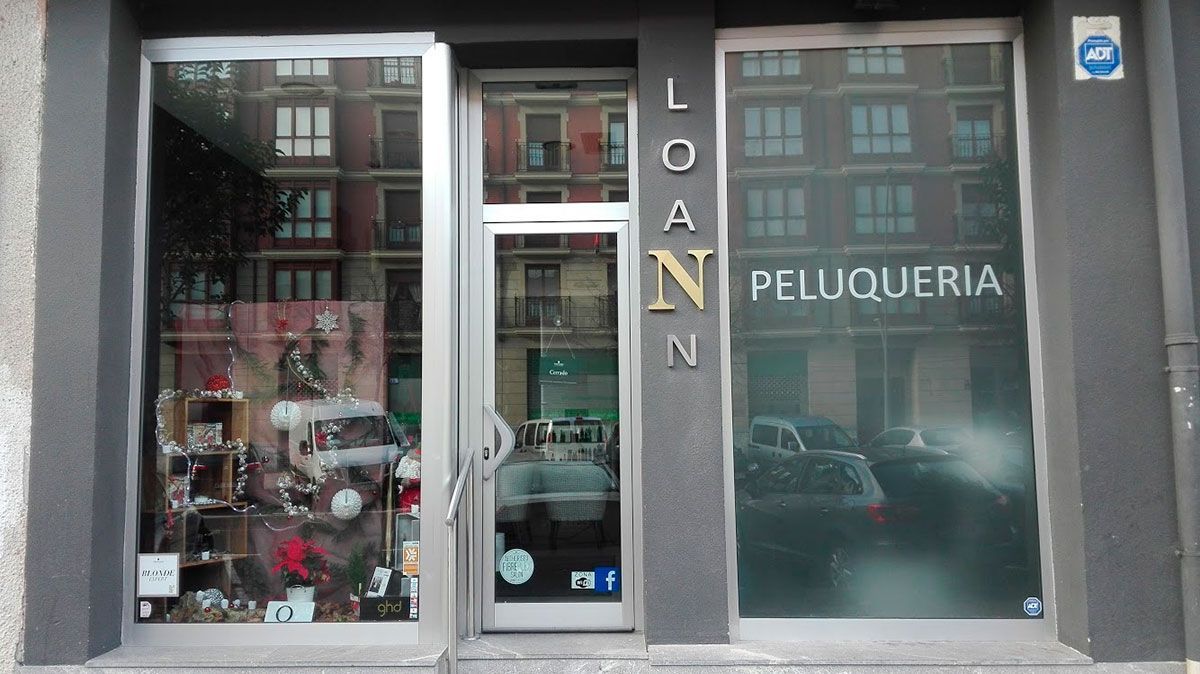 Exterior de una peluquería llamada LOAN PELUQUERIA con reflejos de edificios y coches en las ventanas.