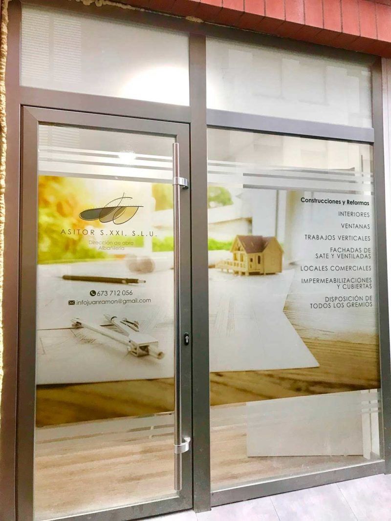 Fachada de tienda con puerta y ventana de cristal, logotipo y texto. Incluye una casa en miniatura, herramientas de diseño y un fondo borroso.