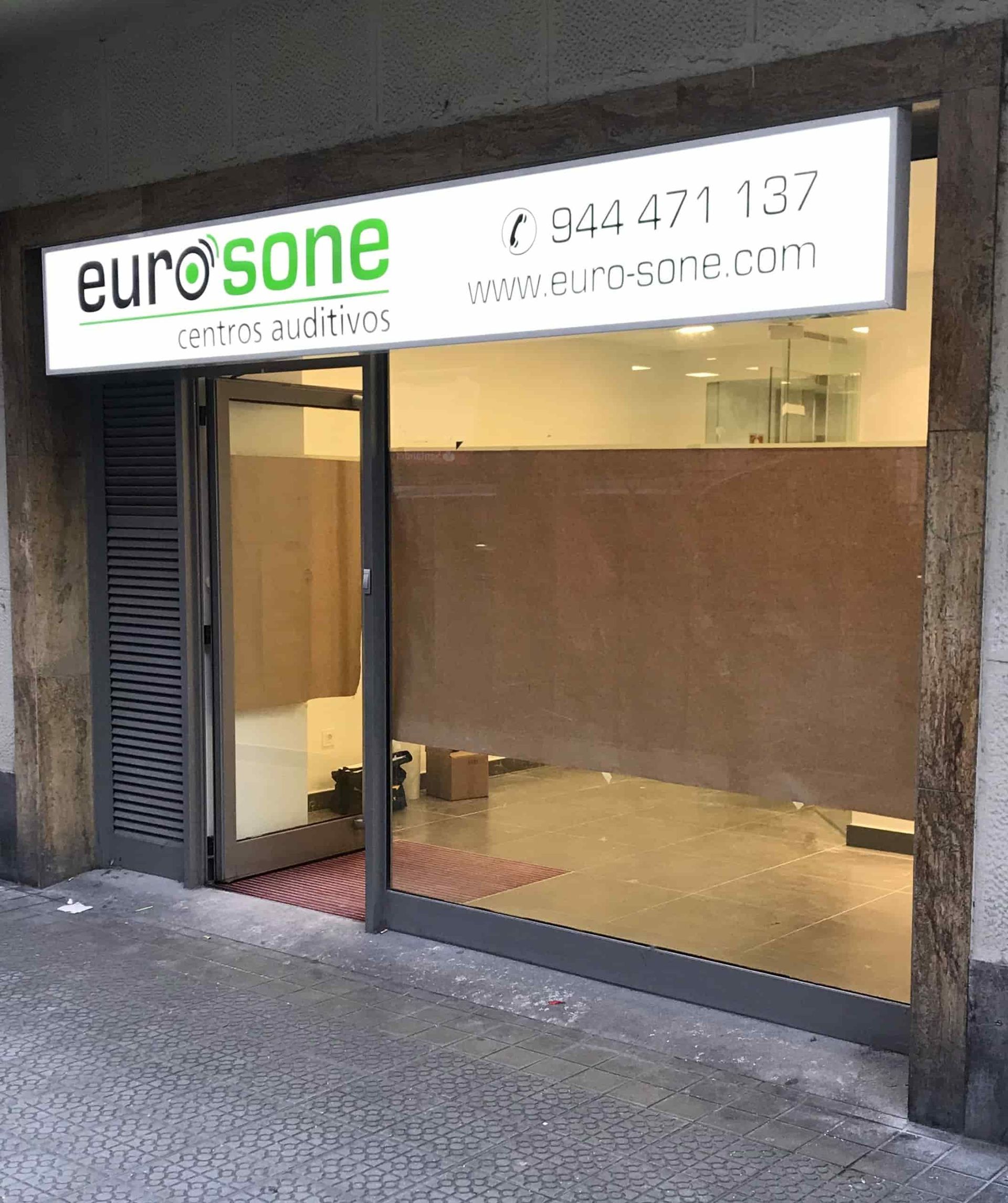 Entrada a EuroSone con puerta de cristal, letrero y número de teléfono. Interior vacío con ventana esmerilada y marco de puerta gris.