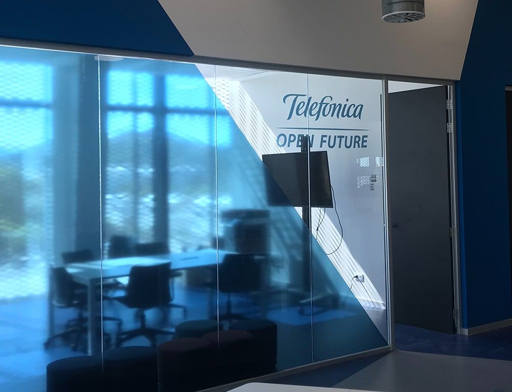 Sala de reuniones de oficina con paredes de cristal tintado azul, logo “Telefonica OPEN FUTURE”, mesa y sillas en su interior.