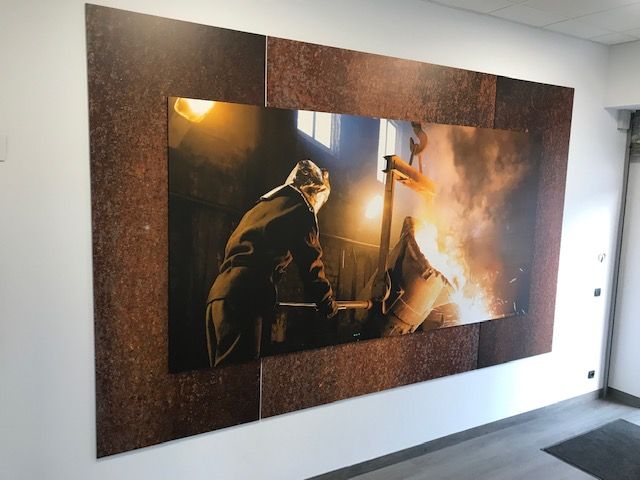 Herrero trabajando cerca de una fragua en llamas. Foto enmarcada en marrón sobre pared blanca.