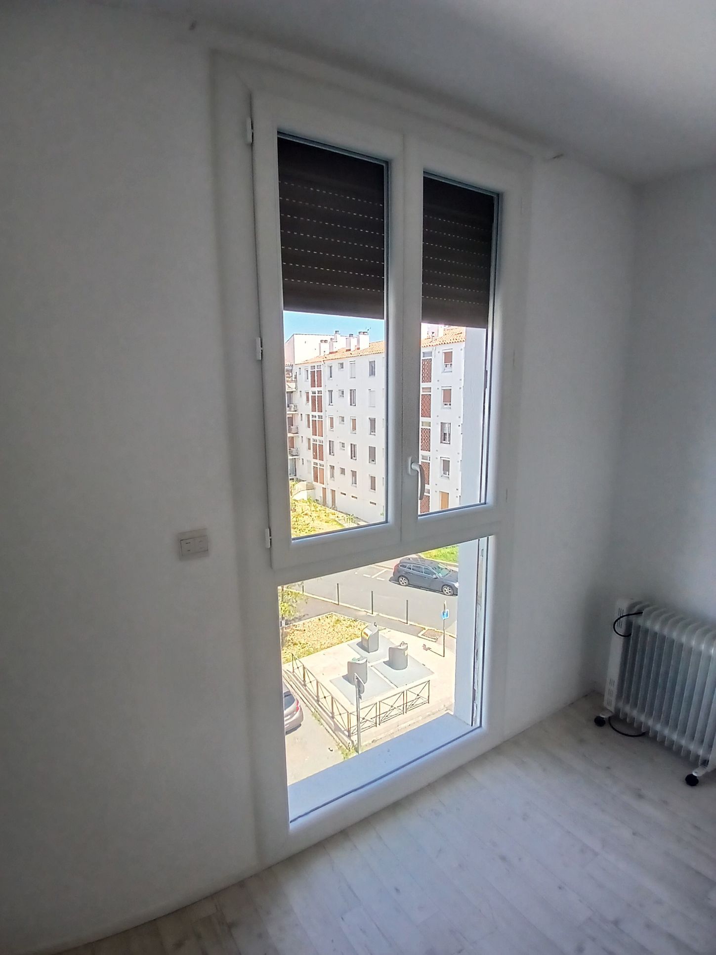 Fenêtre à cadre blanc, stores baissés, donnant sur un paysage urbain. Un radiateur d'appoint est posé à droite.