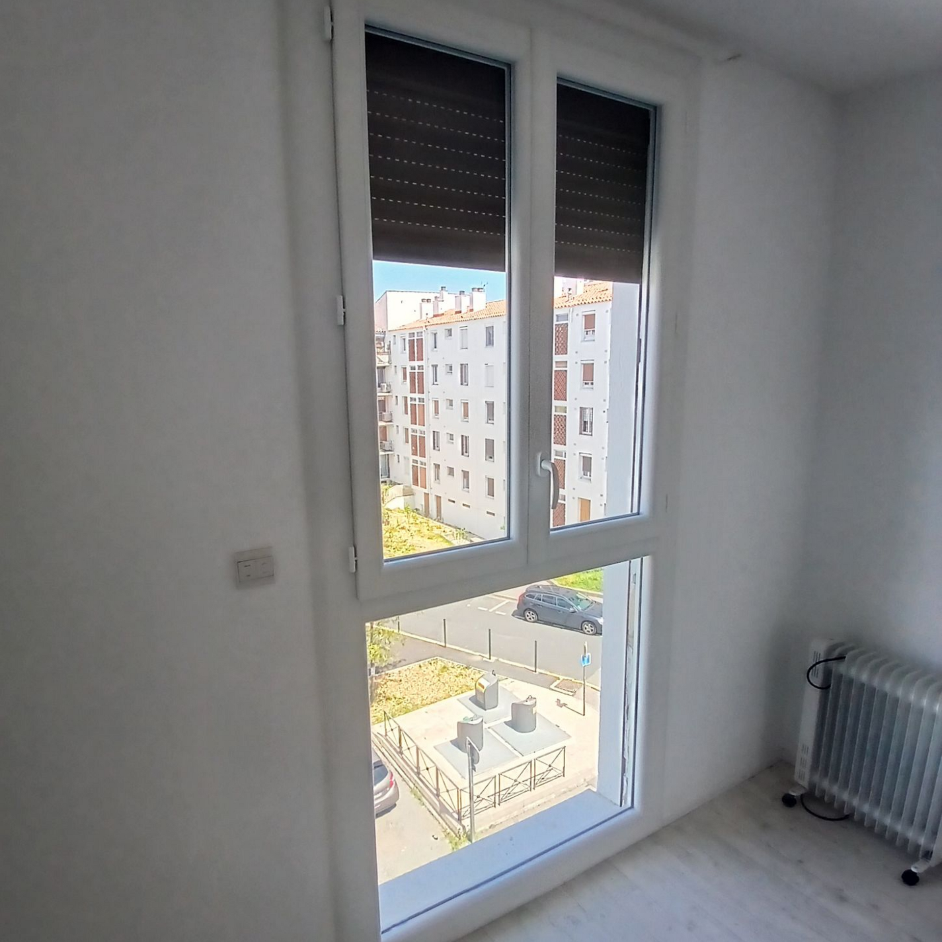 Fenêtre donnant sur un immeuble et une rue. Un radiateur se trouve à droite. La fenêtre est équipée de stores.