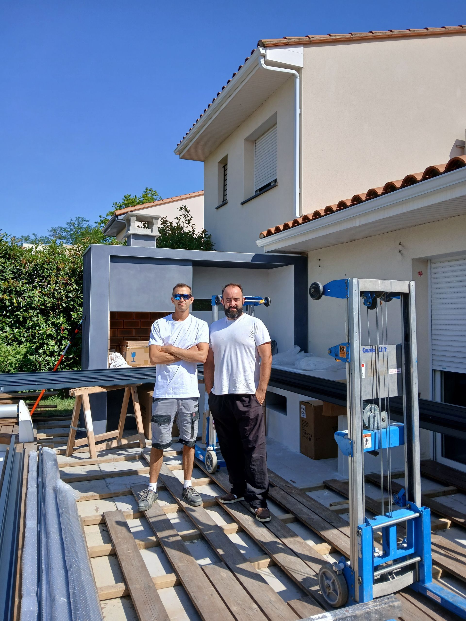 Deux hommes se tiennent sur une terrasse près d'une maison ; on aperçoit des matériaux de construction. L'un d'eux a les bras croisés. Ciel bleu.