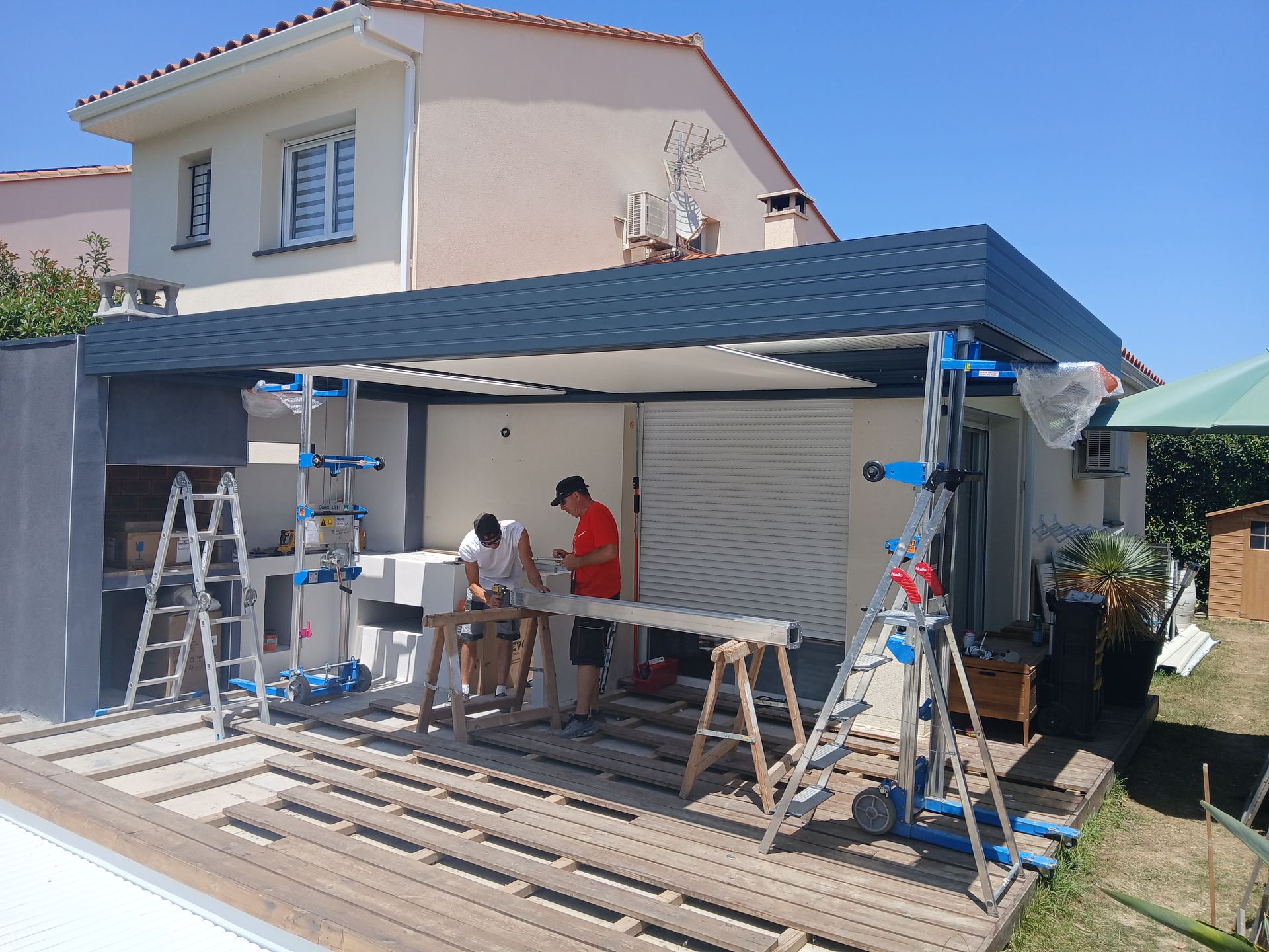 Construction d'une couverture de terrasse : ouvriers installant des poutres sur une terrasse en bois, ciel bleu, maison en arrière-plan.