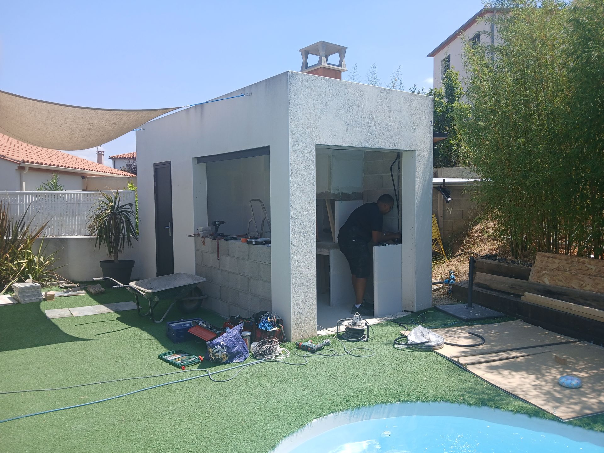 Un homme travaille dans une cuisine extérieure blanche aménagée dans un jardin avec piscine.