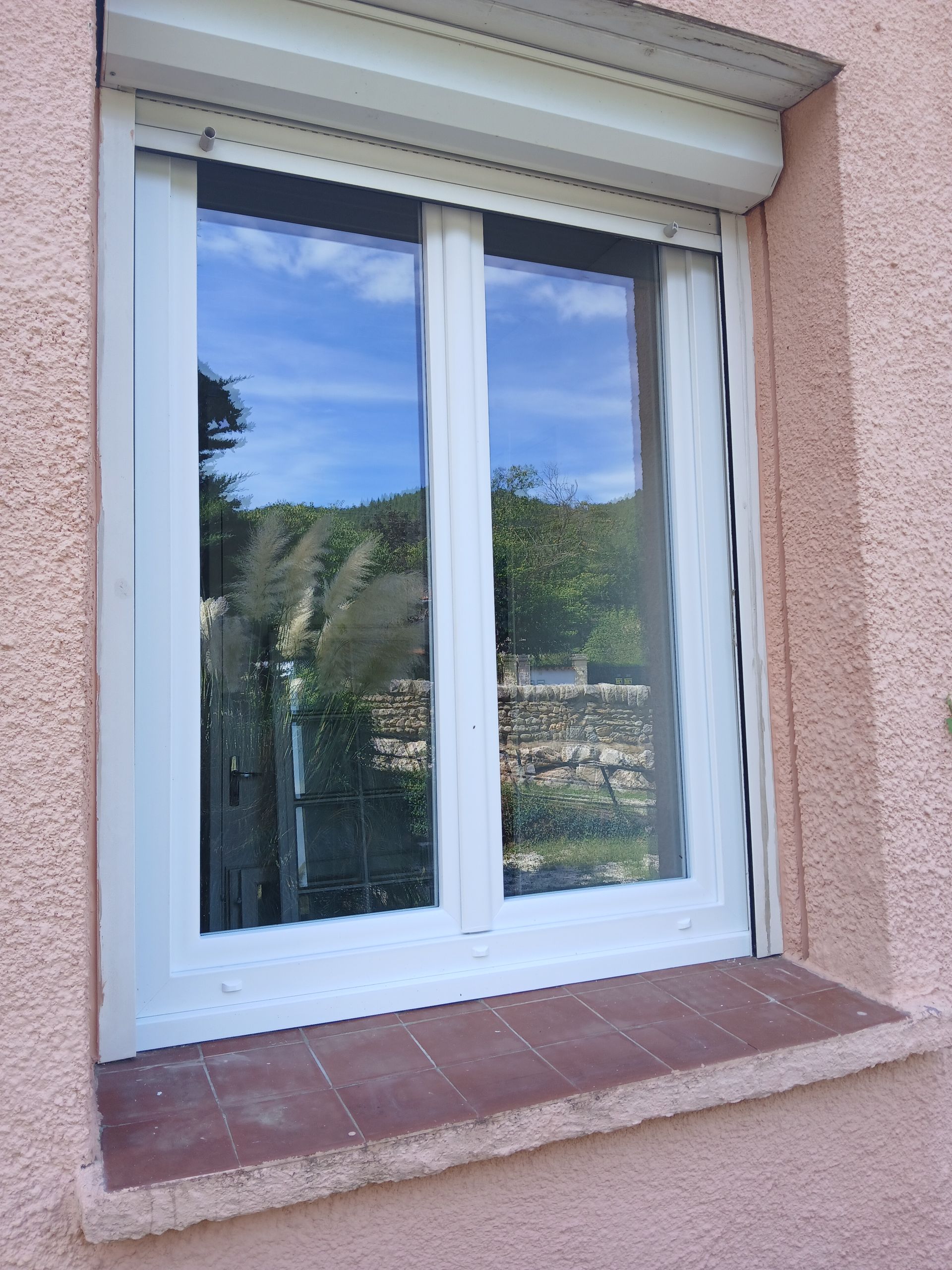 Fenêtre à cadre blanc, volet fermé, reflétant le ciel et la verdure. Mur en stuc rose.