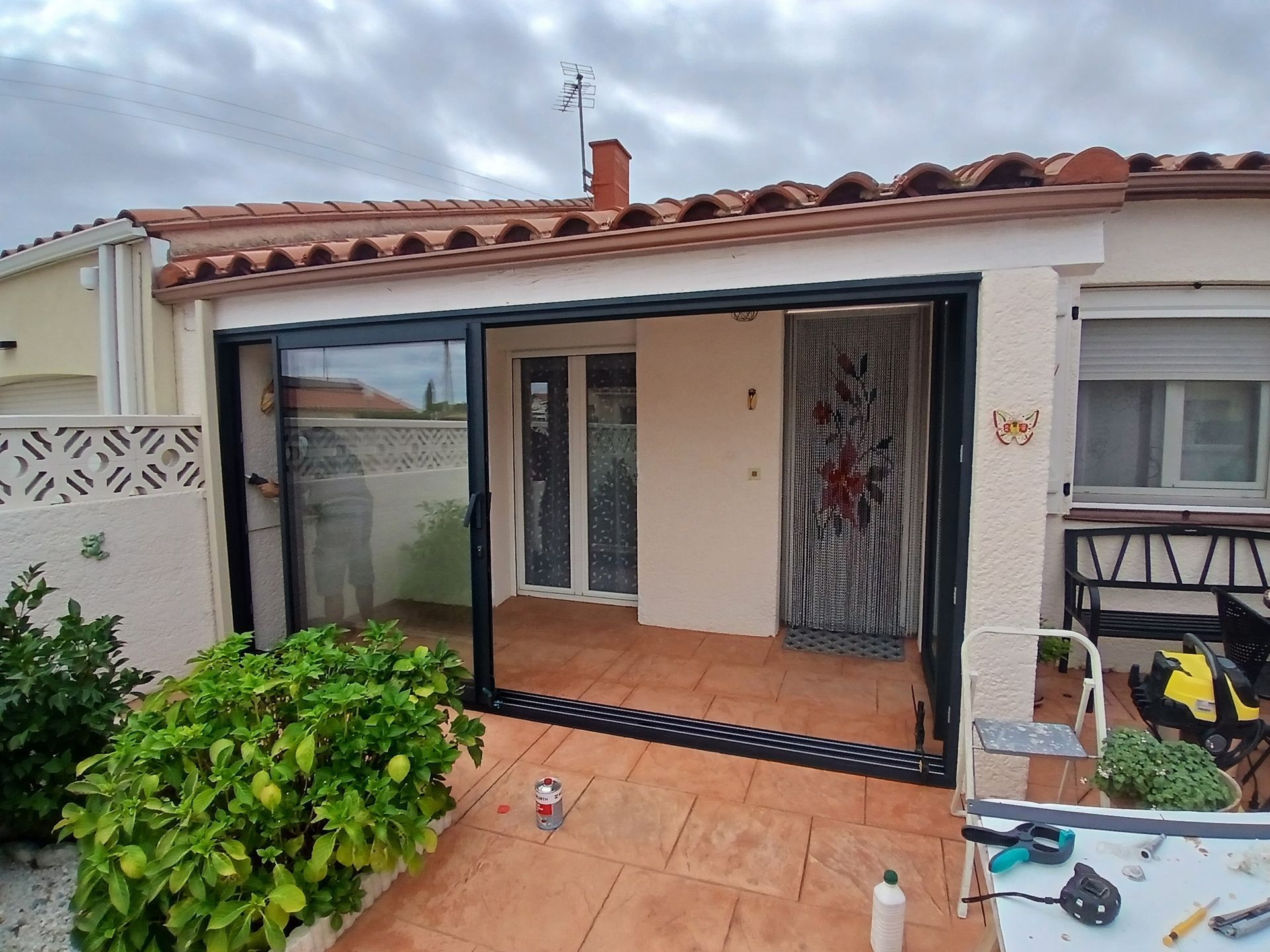 Patio clos avec portes coulissantes en verre à cadre noir. Toit en tuiles rouges et sol en terre cuite. Plantes vertes à l'avant.
