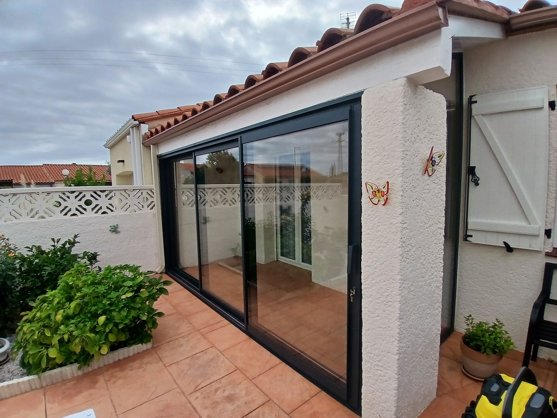 Portes coulissantes en verre donnant sur un patio, sol en carrelage rouge, mur en stuc blanc, plantes et ciel nuageux.