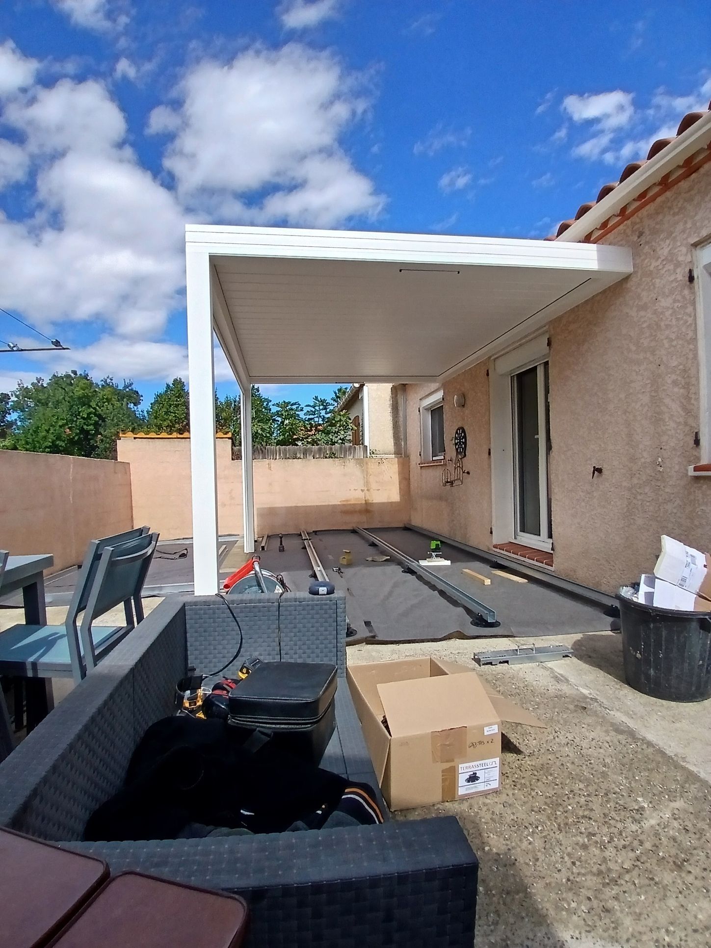 Une pergola blanche, adossée à un bâtiment clair, abrite une terrasse. On aperçoit le ciel bleu, des outils et du mobilier.
