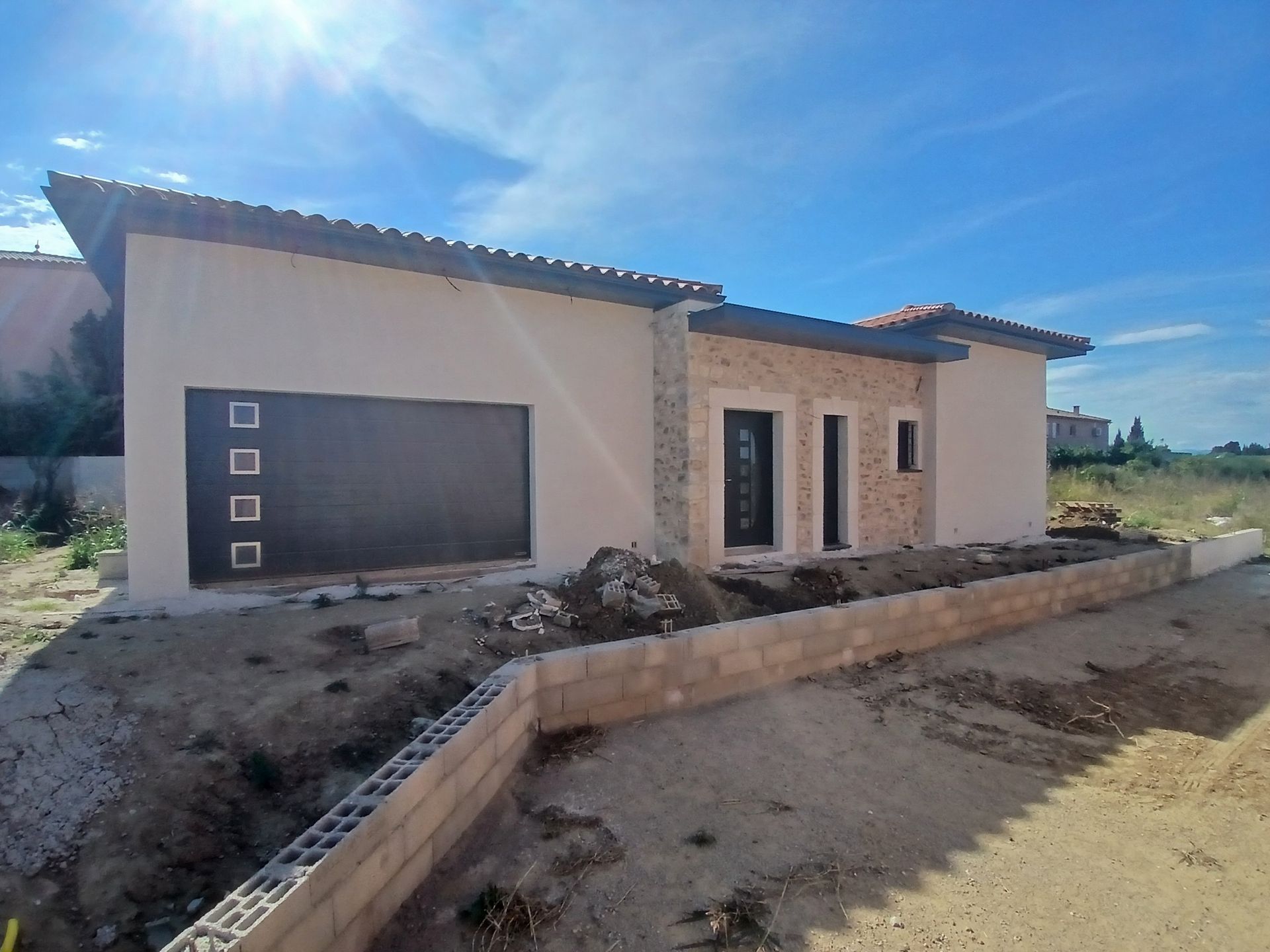 Maison neuve en construction, façade en stuc beige, porte de garage foncée, éléments en pierre, ciel bleu.