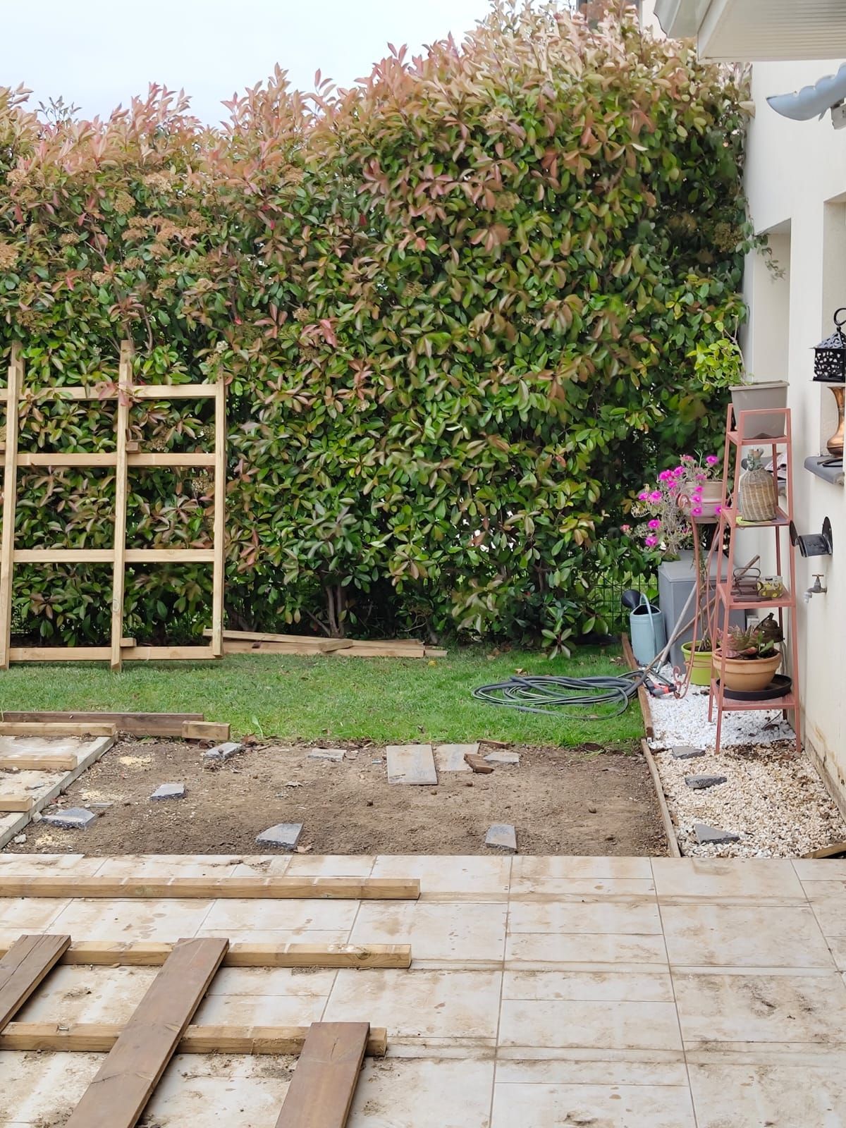 Jardin avec une terrasse en construction et une haie verte. On aperçoit également un treillis en bois et un support pour plantes.