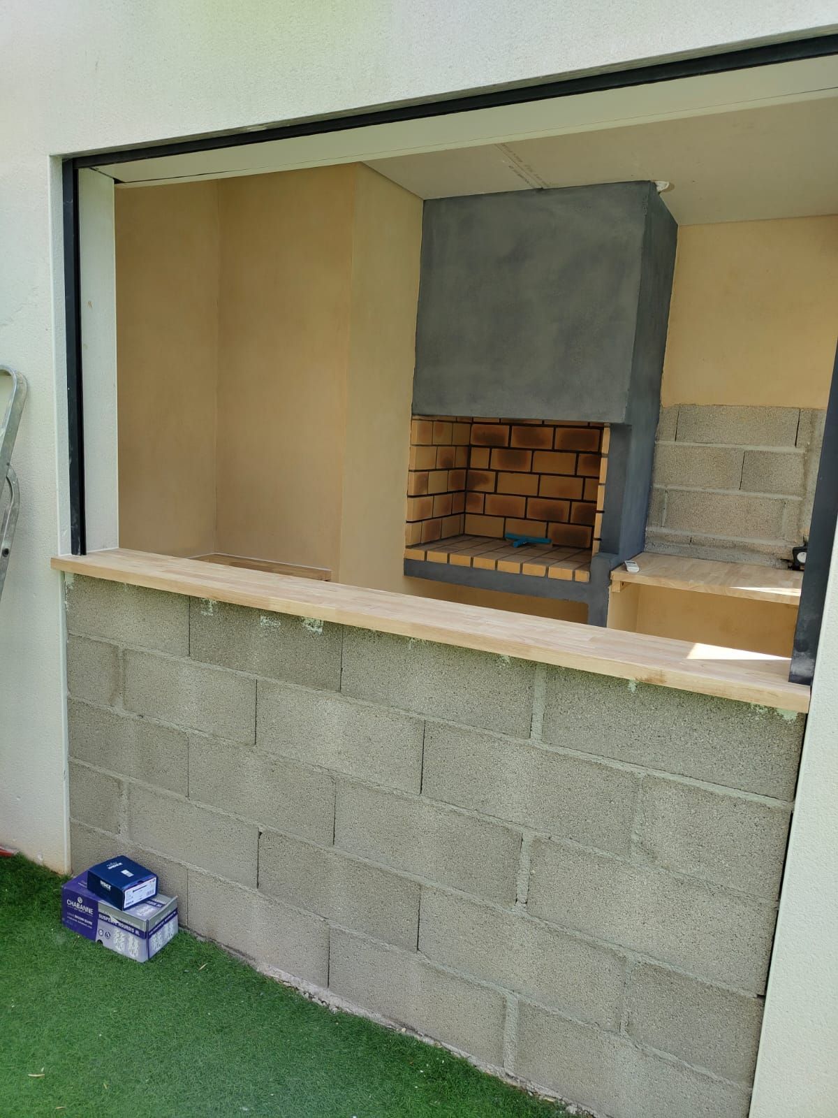 Barbecue extérieur en construction : foyer en briques, comptoir en parpaings gris, ouverture dans le mur, gazon synthétique.