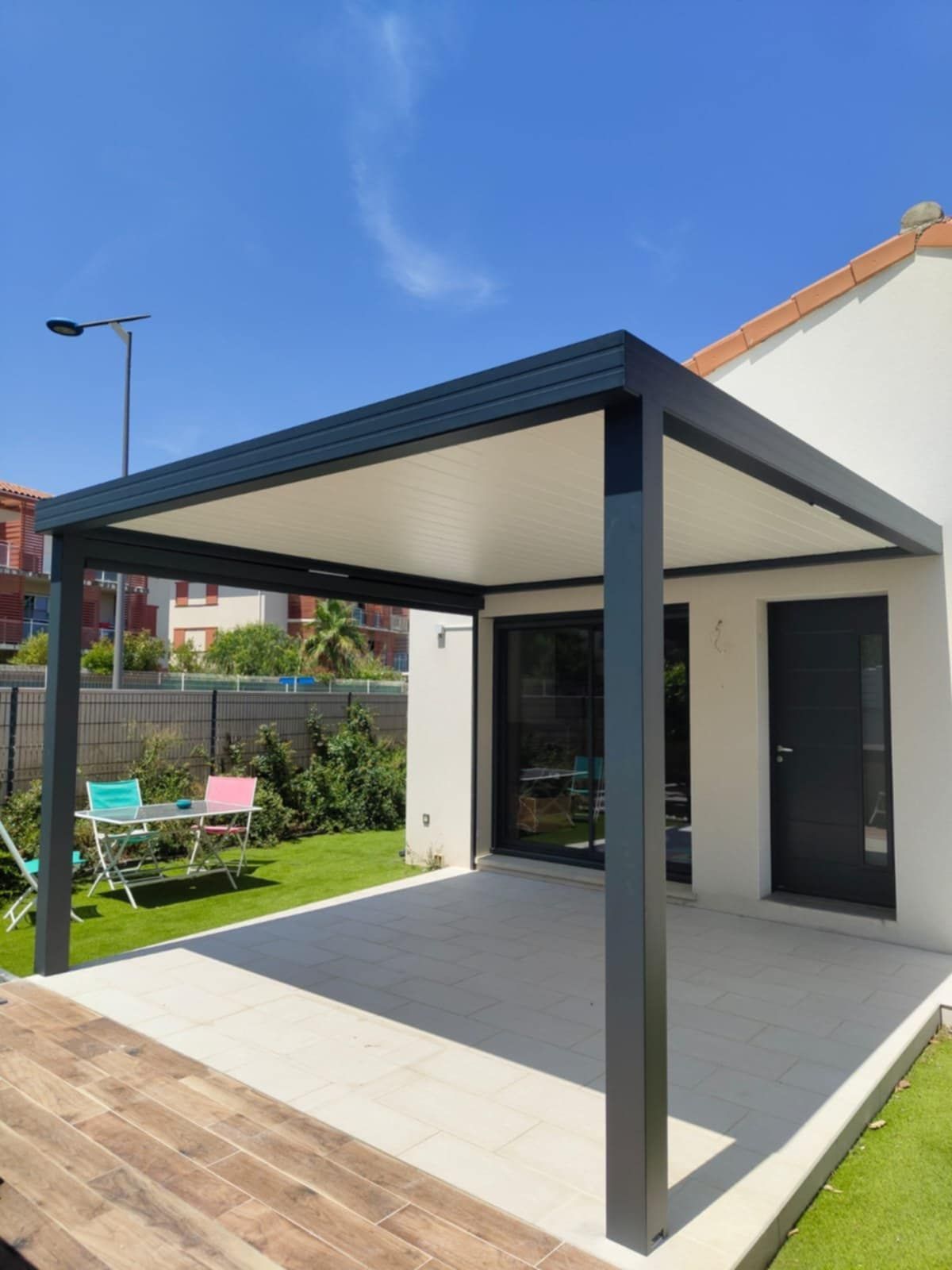 Une pergola moderne à structure sombre surplombe un patio clair, à côté d'une maison sous un ciel bleu.
