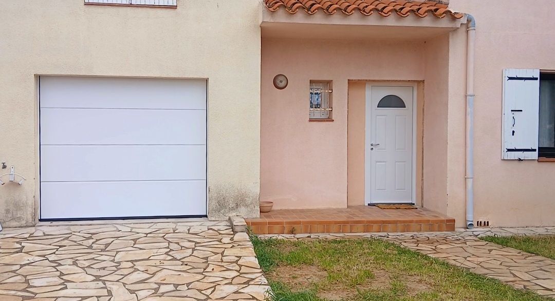 Extérieur d'une maison avec porte de garage et porte d'entrée. Les couleurs claires dominent, avec une allée pavée et de petites zones de gazon.