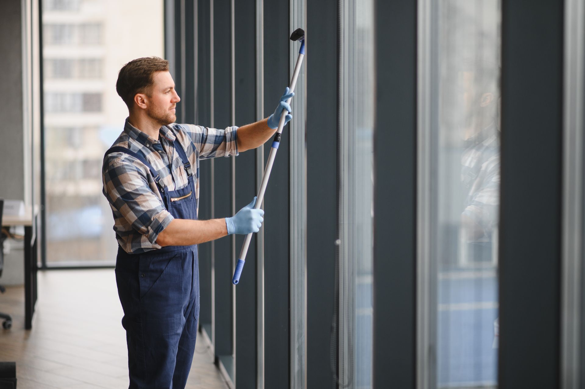 Ein Mann in blauem Overall reinigt hohe Bürofenster mit einem Abzieher mit langem Griff.
