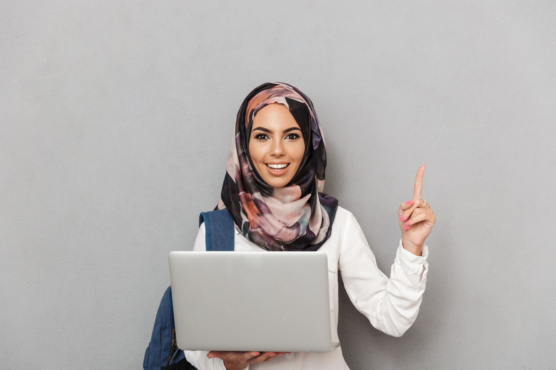 Mujer con hijab sostiene una computadora portátil y señala hacia arriba; fondo gris.