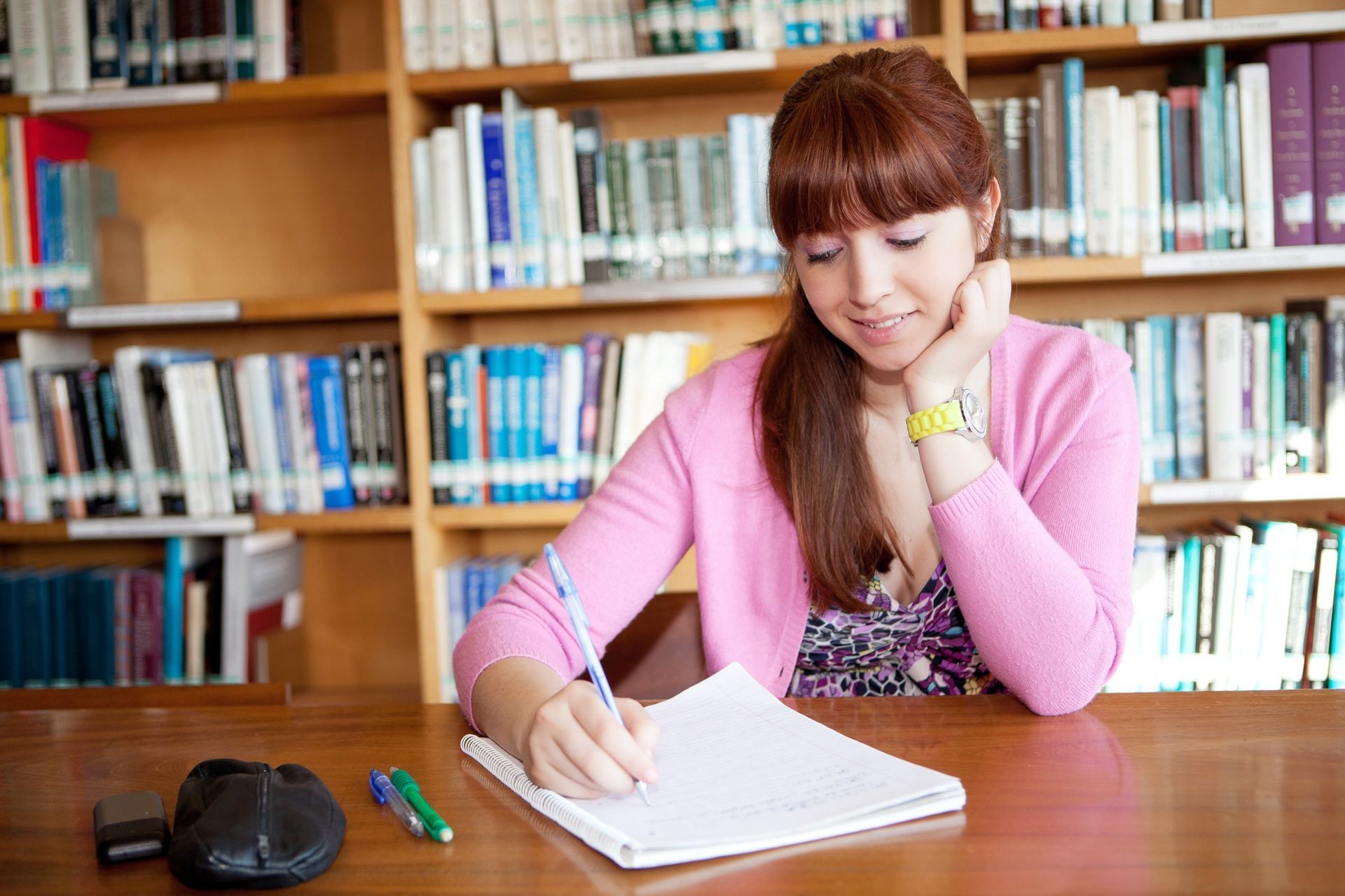Mujer de cabello castaño rojizo escribiendo en un cuaderno en una mesa de biblioteca, sonriendo. Estanterías al fondo.