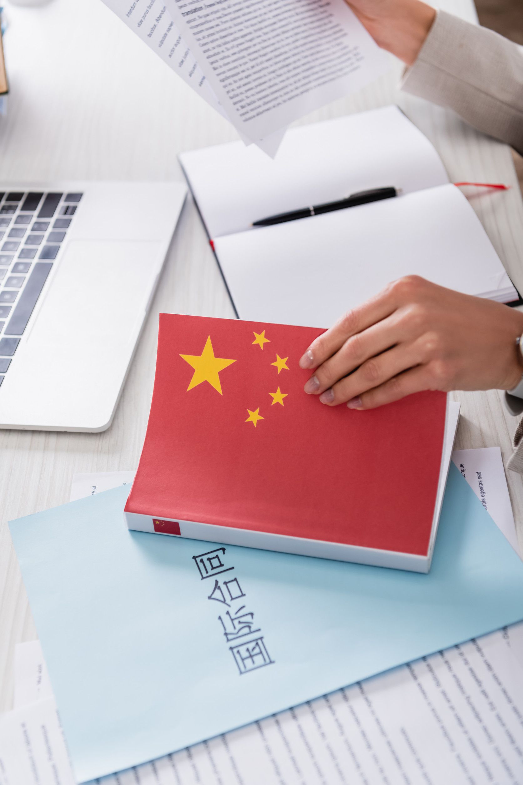 Persona sosteniendo un libro con la bandera china, trabajando en un escritorio con una computadora portátil y papeles.