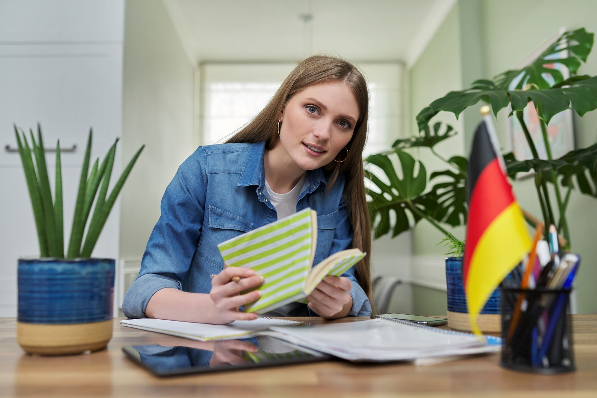 Mujer leyendo un libro en un escritorio con una bandera alemana, plantas y una tableta.