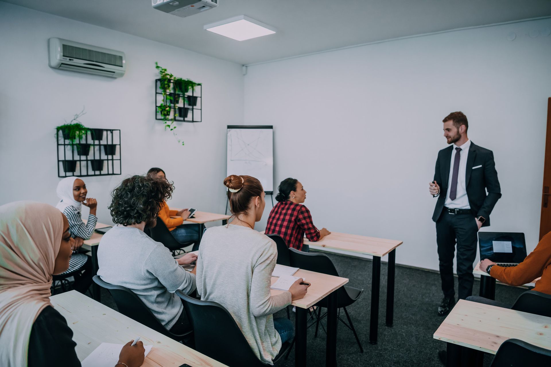 Un hombre de traje da una clase a un grupo diverso en un aula. Los alumnos están sentados en sus escritorios, tomando apuntes.