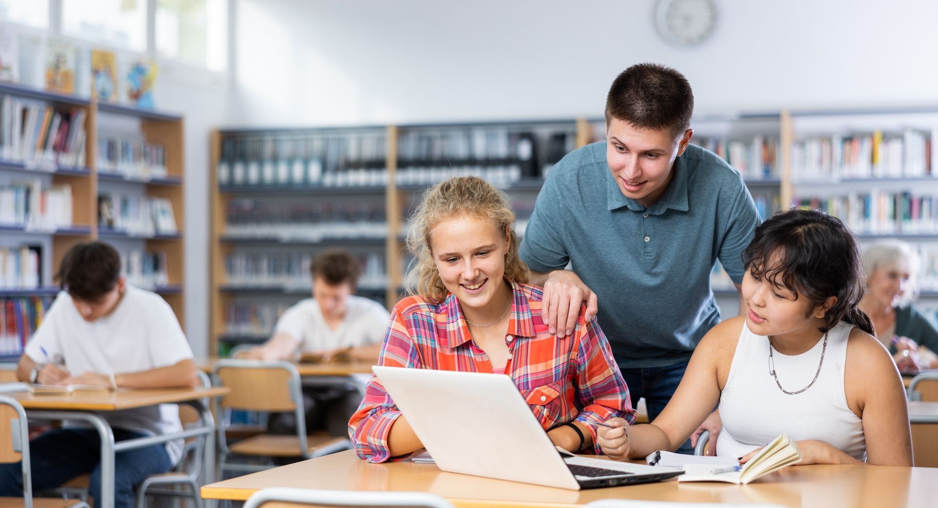 Tres estudiantes sonriendo, mirando una computadora portátil, con un profesor en una biblioteca.