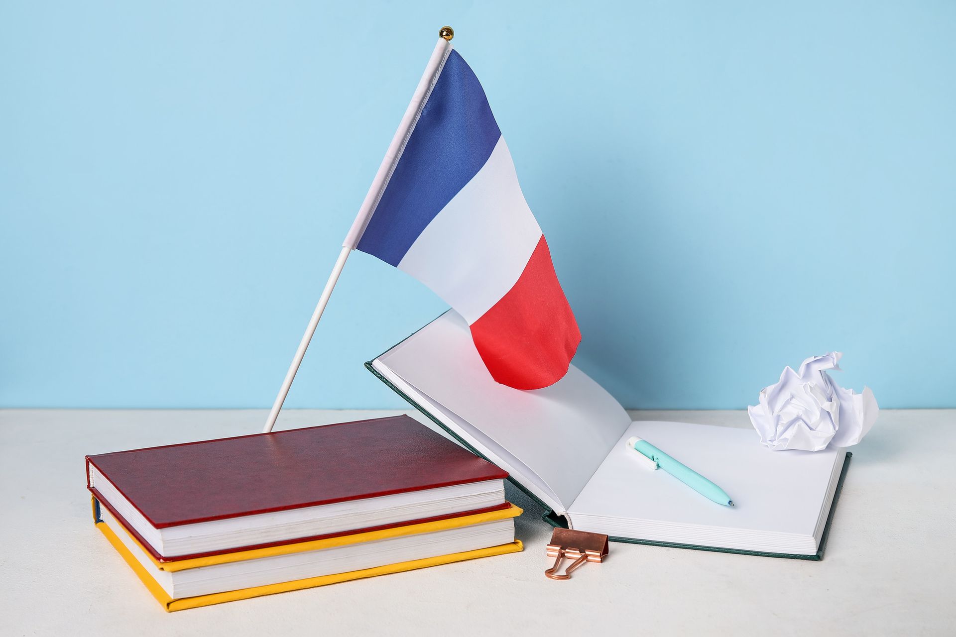 Bandera francesa encima de un cuaderno abierto y libros, con un bolígrafo, papel arrugado, sobre un escritorio.