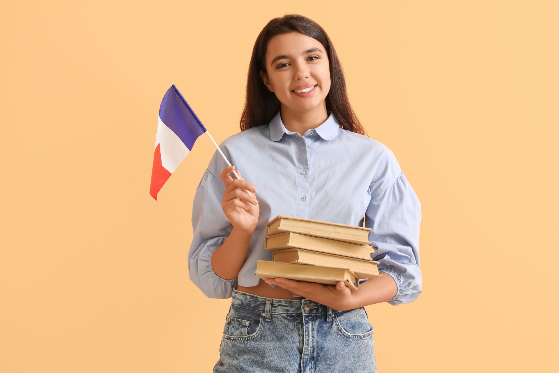 Mujer sosteniendo una bandera francesa y libros, sonriendo, vistiendo una camisa azul y jeans, sobre un fondo color melocotón.